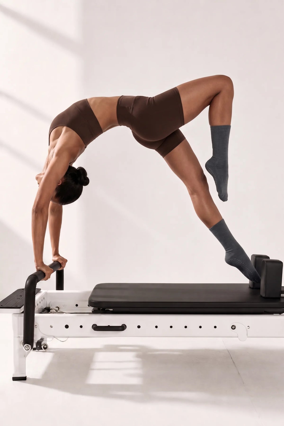 A woman performing an advanced backbend stretch on a Pilates Reformer, wearing YG Socks premium grey crew-length five-toe grip socks for non-slip stability and precise foot alignment on studio equipment.