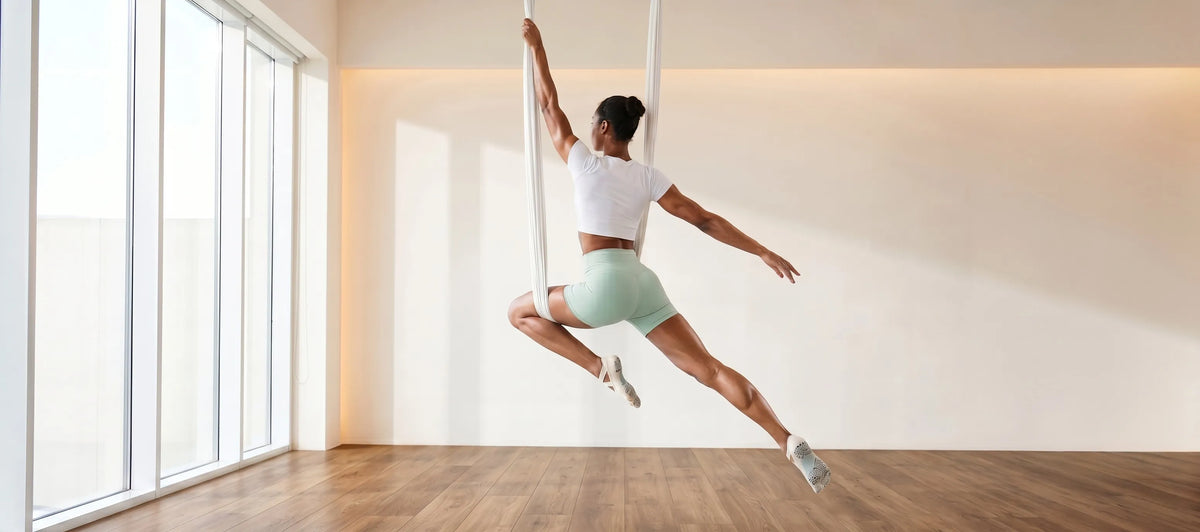 An aerial yoga practitioner elegant in mid-air silks pose, wearing YG Socks premium non-slip five-toe grip socks with grippy soles for ultimate stability and control in a sunlit studio.