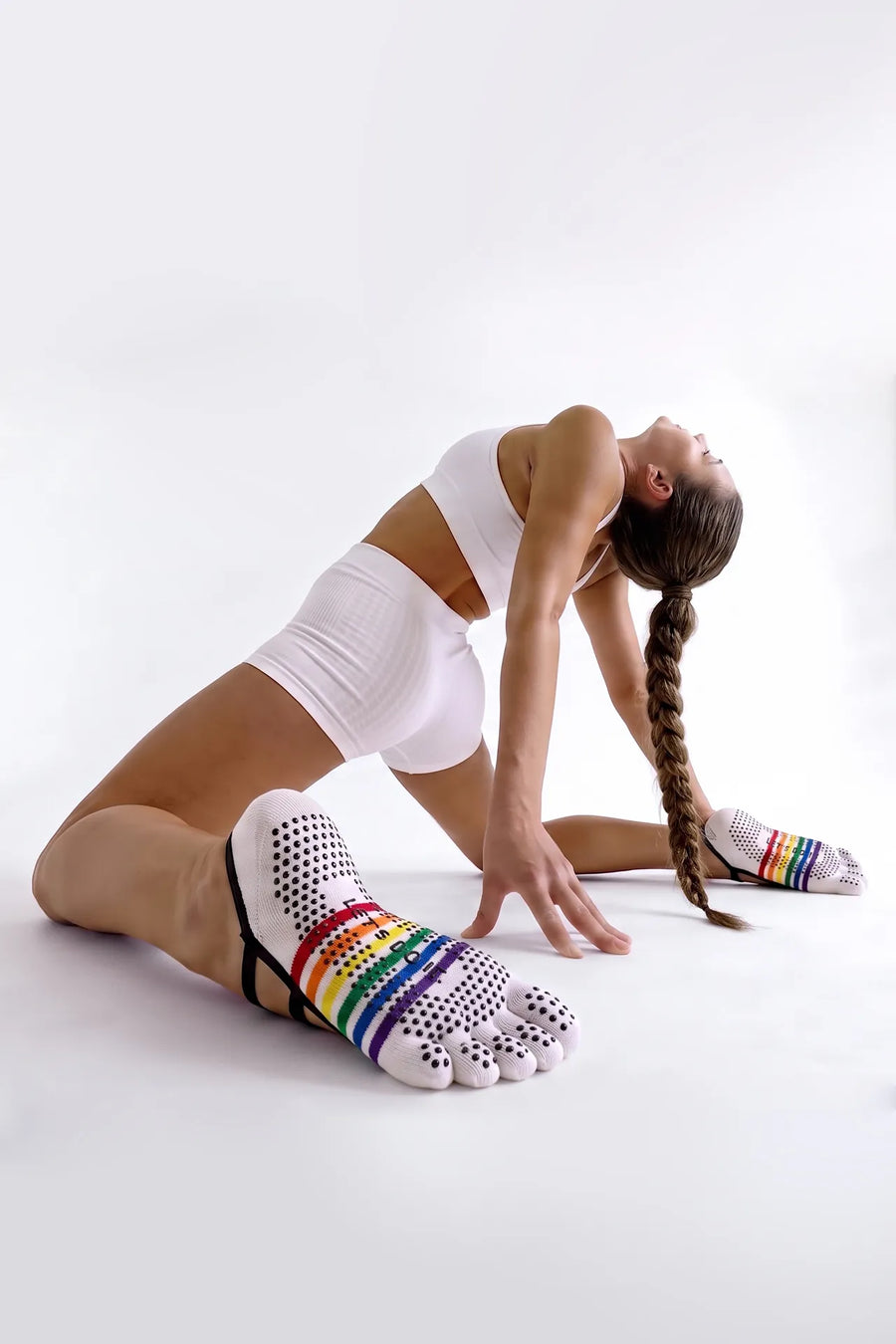 A woman with a long braid, wearing a white sports bra and shorts, performs a deep backbend stretch on a white studio floor. In the foreground, a close-up view of her foot in a white five-toe grip sock with a colorful rainbow stripe and silicone dots is prominently displayed. The background is a plain, seamless white void, emphasizing the figure and the details of the sock.