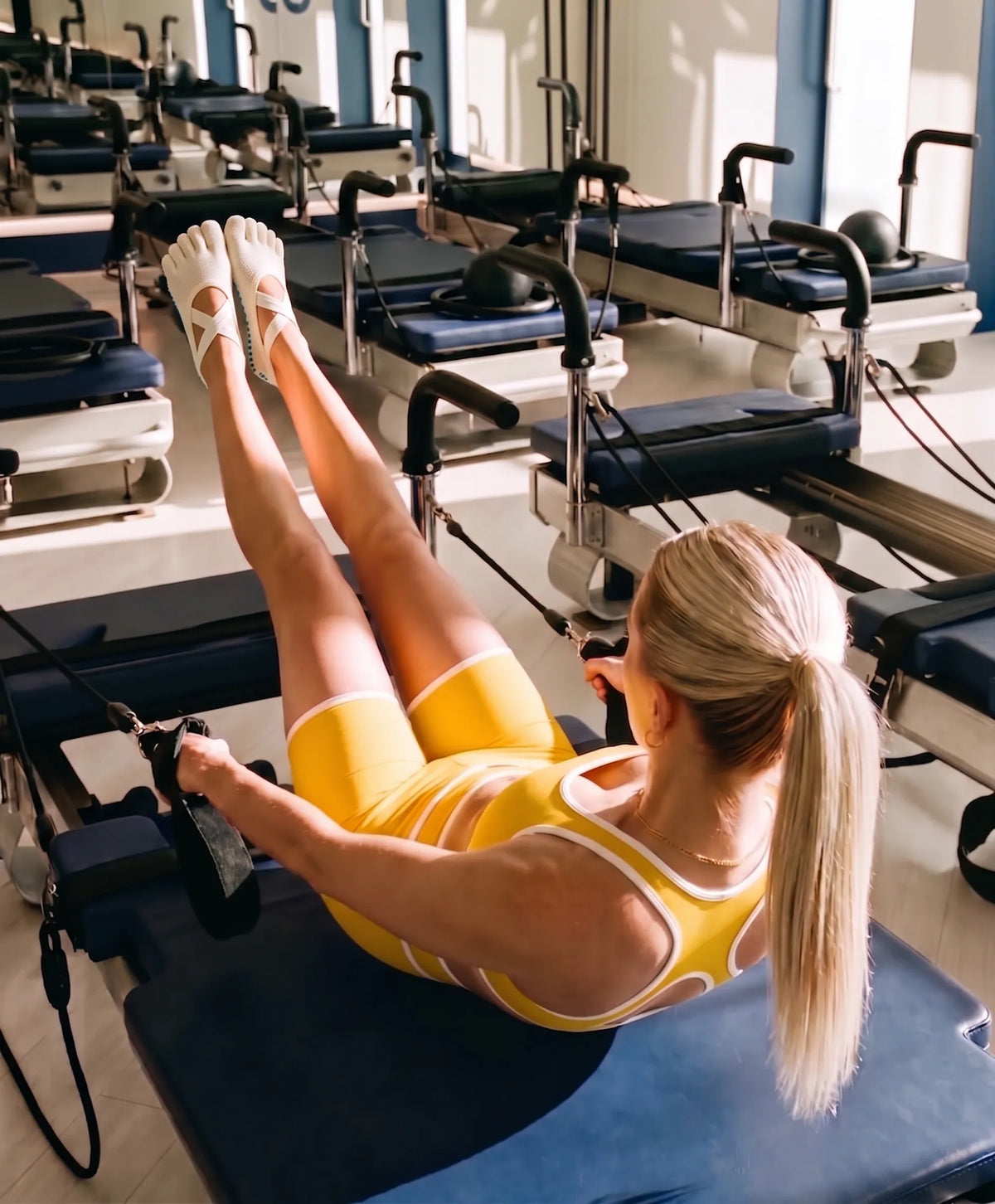 A woman with a high ponytail, wearing a yellow sports bra and matching shorts, lies supine on a blue Pilates reformer machine. She holds the reformer straps with her hands, performing an exercise with her legs extended upwards and pointed towards the ceiling. Her feet are clad in white, five-toe grip socks, providing stability on the machine. The background features a row of identical blue Pilates reformers in a well-lit studio, with reflections visible in a mirror. 
